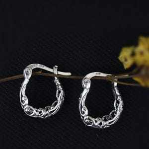 May include: A pair of silver hoop earrings with an intricate, ornate design. The earrings are delicate and have a slightly curved shape.