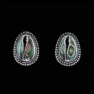 Puede incluir: Un par de pendientes de botón ovalados. Cada pendiente presenta un borde plateado en forma de cuerda que rodea una incrustación de concha de abulón. La concha de abulón muestra colores iridiscentes de verde, azul y morado. Los pendientes están sobre un fondo negro.