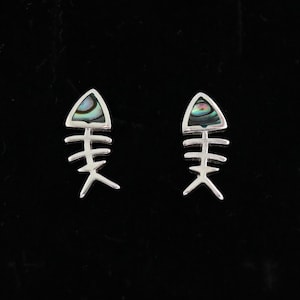 Pode incluir: Um par de brincos de pino prateados em forma de espinhas de peixe. Cada brinco apresenta uma cabeça triangular incrustada com um segmento de concha de abalone polido e iridescente que exibe tons rodopiantes de verde, azul e roxo no metal.