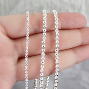 May include: Close-up of three silver ball chains of varying sizes. The chains are held in a hand against a soft, blurred background. The chains are made of small, round, shiny beads, reflecting light.