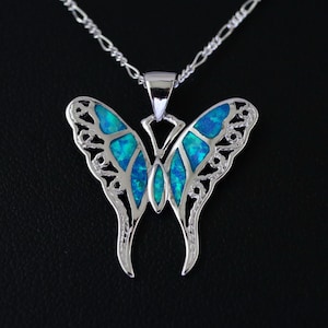 Könnte beinhalten: Eine silberne Schmetterlings-Anhänger-Halskette mit blauem Opal-Inlay. Das Schmetterlingsdesign weist filigrane Ausschnitte an den Flügeln und eine zarte Kette auf. Der Anhänger ist vor einem schwarzen Hintergrund zentriert.