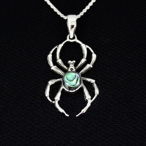 Puede incluir: Un collar con colgante de araña plateada con un centro de concha de abulón. La araña tiene ocho patas y está suspendida de una delicada cadena plateada. El colgante está sobre un fondo negro.