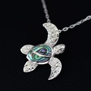 May include: A silver turtle pendant necklace with a chain. The turtle pendant has an iridescent shell in shades of blue, green, and purple. The necklace is shown against a black backdrop.