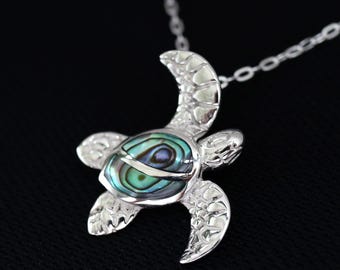May include: A silver turtle pendant necklace with a chain. The turtle pendant has an iridescent shell in shades of blue, green, and purple. The necklace is shown against a black backdrop.