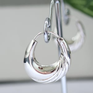 Peut inclure: Une paire de boucles d'oreilles créoles en argent poli. Les boucles d'oreilles ont une forme arrondie en croissant avec un motif texturé et nervuré sur la surface extérieure. Elles sont présentées sur un présentoir à boucles d'oreilles argenté.