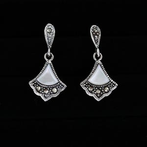 May include: A pair of silver-toned earrings with a fan-shaped design. Each earring features a white, pearlescent center, surrounded by a detailed silver border with small, dark accents. The earrings hang from a decorative top piece.
