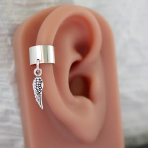 May include: Silver-colored ear cuff with a dangling wing charm. The cuff wraps around the ear's cartilage, and the wing charm hangs below. The wing has detailed feather etching.