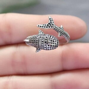 K&ouml;nnte beinhalten: Ein silberfarbener Ring in Form eines Walhais. Der Ring hat ein detailliertes Design mit einem strukturierten K&ouml;rper und einem Schwanz, der sich um den Finger wickelt. Der K&ouml;rper des Walhais ist mit kleinen, dunklen Punkten bedeckt.