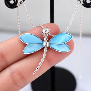 May include: A silver-toned dragonfly pendant necklace featuring four iridescent light blue wings and a segmented tail. The pendant is suspended from a delicate silver-toned chain, showcasing a polished metallic finish and smooth, vibrant blue inlay.