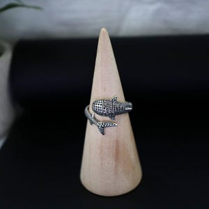 K&ouml;nnte beinhalten: Ein silberner Ring in Form eines Walhais. Der Ring umschlie&szlig;t den Finger, wobei der K&ouml;rper und die Schwanzflosse des Hais das Design bilden. Der K&ouml;rper des Hais ist mit kleinen, dunklen Punkten versehen.