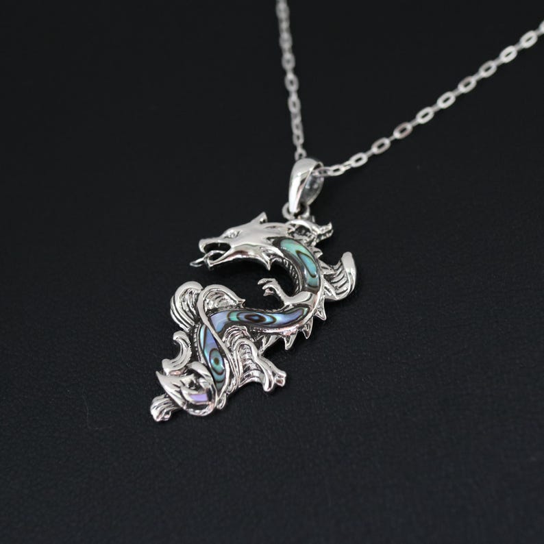 May include: A silver dragon pendant necklace with iridescent abalone shell inlay. The dragon is detailed with scales and a fierce expression, suspended from a delicate chain. The pendant is approximately 2 inches in length.