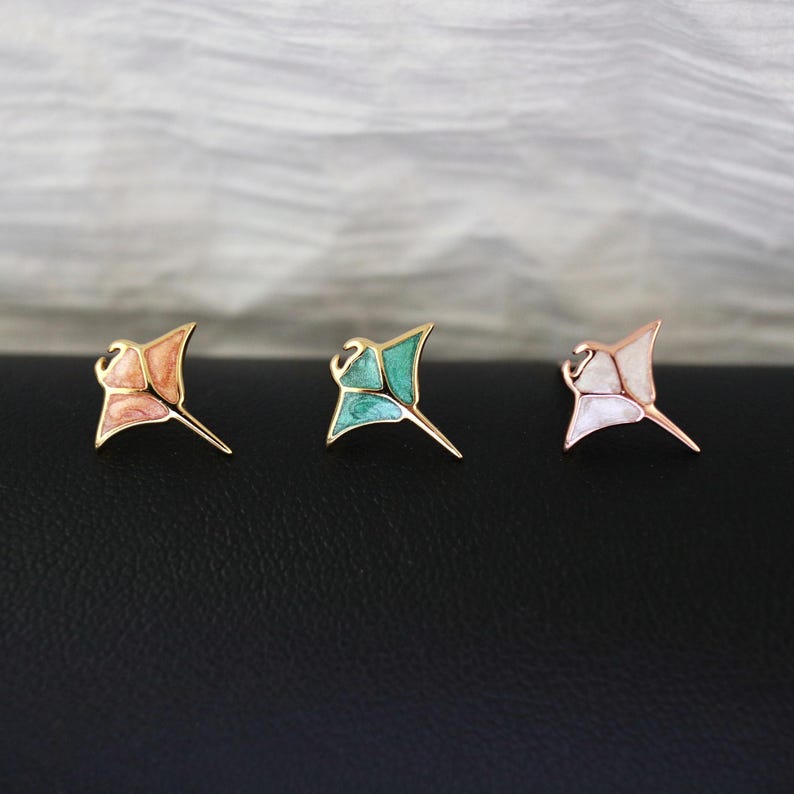May include: Three stingray-shaped stud earrings with gold-toned metal outlines. The left earring features a peach-colored enamel inlay, the center earring has a teal enamel inlay, and the right earring contains a white pearlescent enamel inlay.
