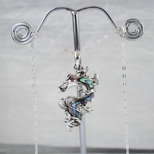May include: A silver dragon pendant necklace with iridescent abalone shell inlay. The dragon is detailed with scales and a flowing design, suspended from a delicate silver chain. The pendant is displayed on a silver jewelry stand.