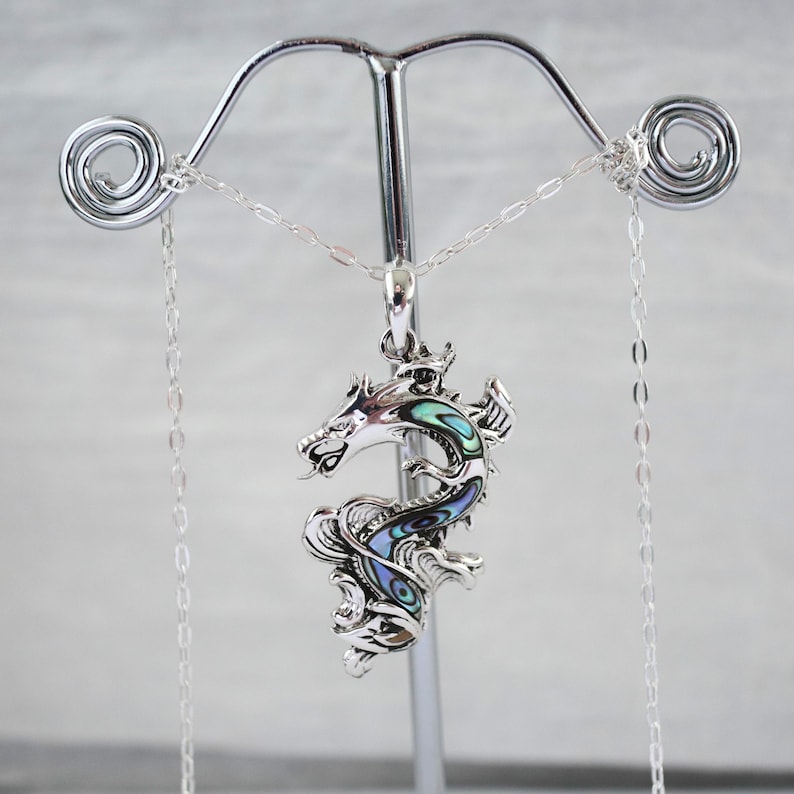 May include: A silver dragon pendant necklace with iridescent abalone shell inlay. The dragon is detailed with scales and a flowing design, suspended from a delicate silver chain. The pendant is displayed on a silver jewelry stand.