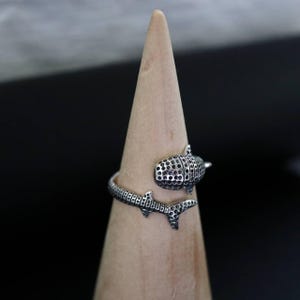 K&ouml;nnte beinhalten: Ein silberner Ring mit einem Walhai-Design. Der Ring umschlie&szlig;t den Finger, wobei der K&ouml;rper und die Schwanzflosse des Walhais das Band bilden. Der K&ouml;rper des Walhais hat ein strukturiertes Muster mit kleinen, dunklen L&ouml;chern.
