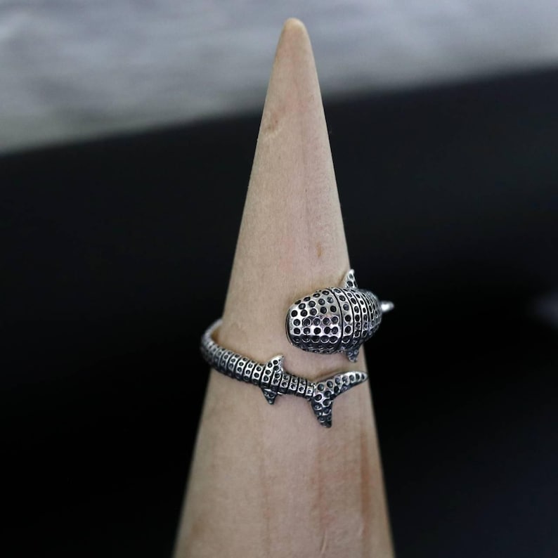 K&ouml;nnte beinhalten: Ein silberner Ring mit einem Walhai-Design. Der Ring umschlie&szlig;t den Finger, wobei der K&ouml;rper und die Schwanzflosse des Walhais das Band bilden. Der K&ouml;rper des Walhais hat ein strukturiertes Muster mit kleinen, dunklen L&ouml;chern.