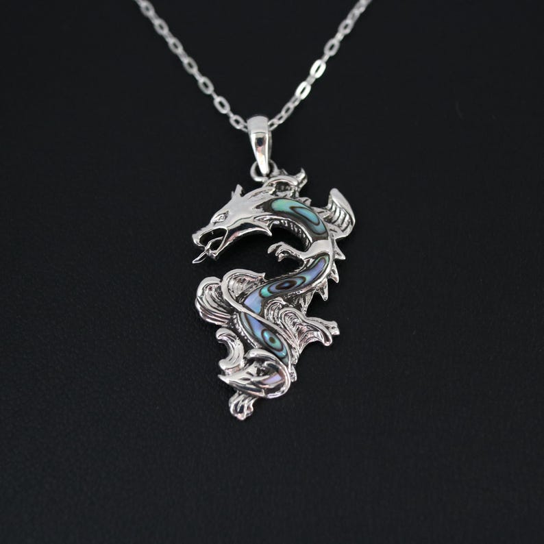 May include: A silver dragon pendant with a detailed design and a shimmering abalone inlay in shades of blue, green, and purple. The dragon is suspended from a delicate silver chain, creating a striking piece of jewelry.
