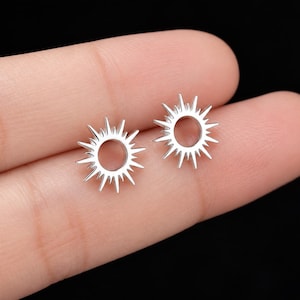 May include: A pair of silver-toned stud earrings shaped like sunbursts with a circular center and radiating pointed rays. The earrings feature a polished metallic finish and a simple, open design that highlights the star-like geometric pattern.