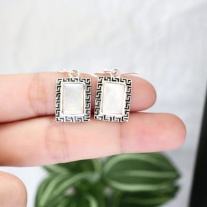 May include: A pair of rectangular silver earrings with a white, iridescent center stone. The silver frame features a black Greek key design. The earrings are shown against a blurred background.
