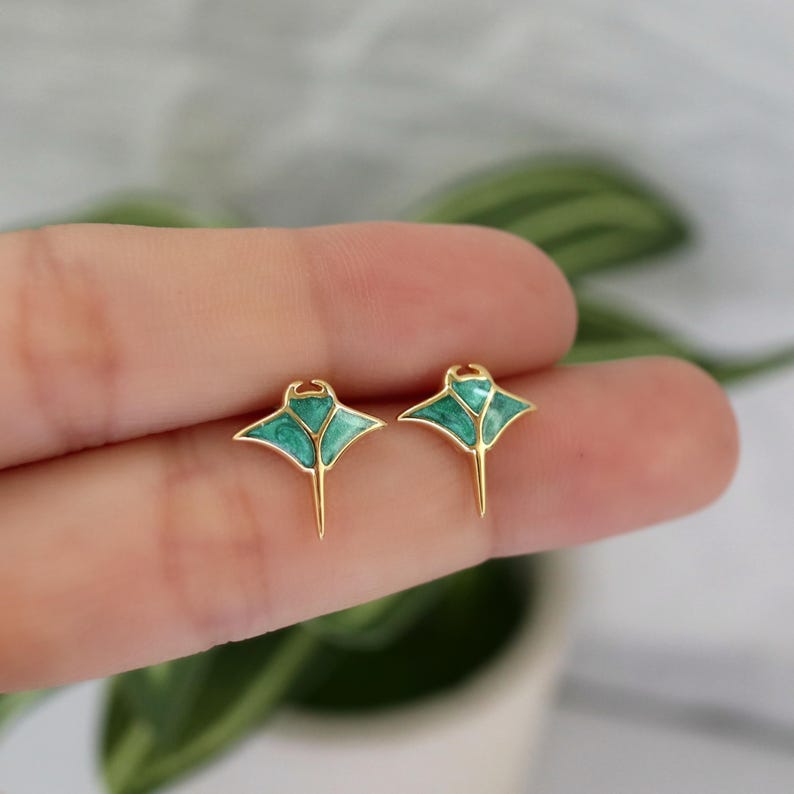 May include: A pair of gold-toned stud earrings shaped like manta rays, featuring a vibrant teal and green marbled enamel inlay within the metal outlines. The earrings have a pointed tail and wide, triangular wings, creating a distinct aquatic silhouette.