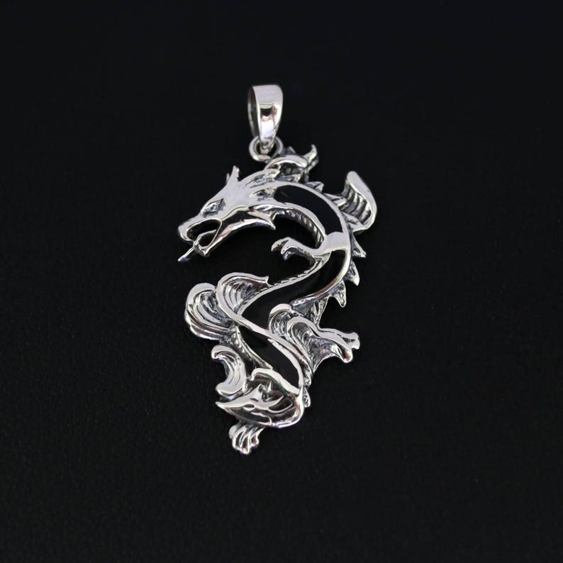 May include: A silver dragon pendant with intricate detailing. The dragon is depicted in a dynamic pose, with its body curving and its mouth open. The pendant has a loop at the top for a chain. The background is black.