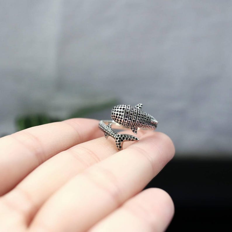 K&ouml;nnte beinhalten: Ein silberner Ring mit einem Walhai-Design. Der Ring umschlie&szlig;t den Finger, wobei der K&ouml;rper und die Schwanzflosse des Walhais das Design bilden. Der K&ouml;rper des Walhais ist mit kleinen, dunklen, kreisf&ouml;rmigen Mustern versehen.