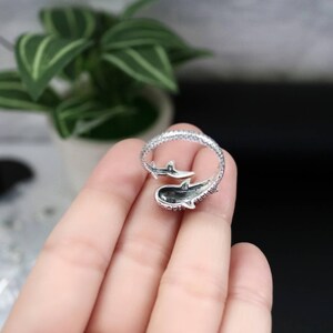 K&ouml;nnte beinhalten: Ein silberner Ring mit einem Walhai-Design. Der Ring umschlie&szlig;t den Finger, wobei der K&ouml;rper des Hais das Hauptelement bildet. Der Ring ist mit kleinen Verzierungen versehen. Der Hintergrund ist verschwommen, mit einer Pflanze sichtbar.