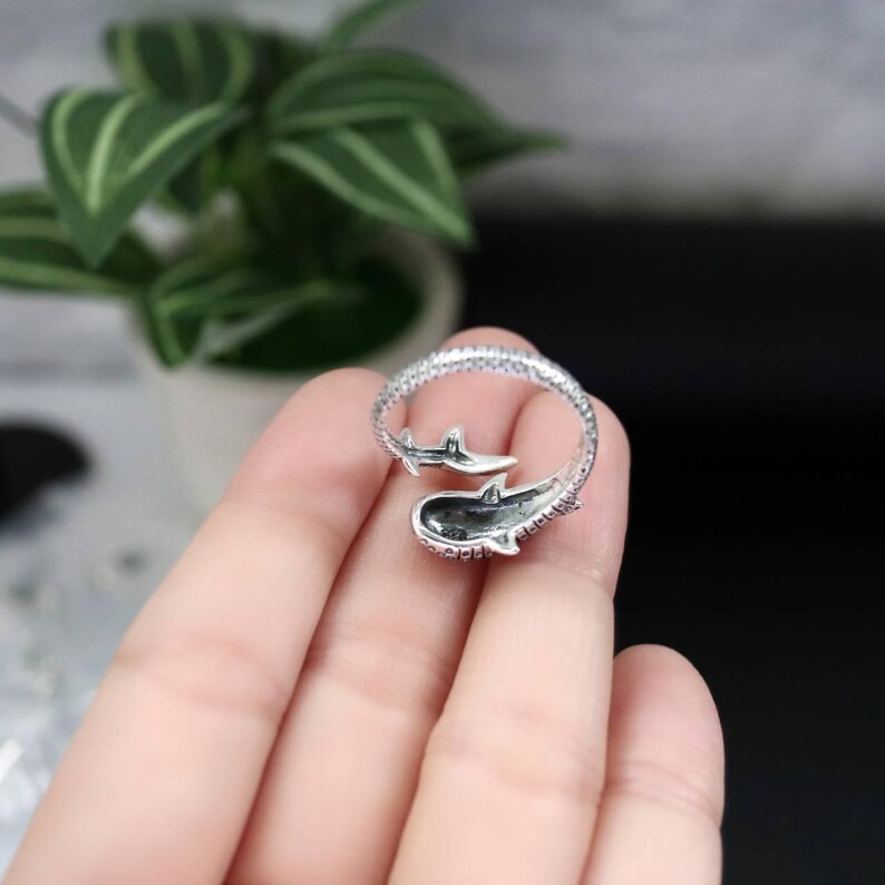 K&ouml;nnte beinhalten: Ein silberner Ring mit einem Walhai-Design. Der Ring umschlie&szlig;t den Finger, wobei der K&ouml;rper des Hais das Hauptelement bildet. Der Ring ist mit kleinen Verzierungen versehen. Der Hintergrund ist verschwommen, mit einer Pflanze sichtbar.