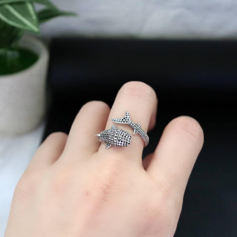 K&ouml;nnte beinhalten: Ein silberner Ring mit einem Walhai-Design. Der Ring umschlie&szlig;t den Finger, wobei der K&ouml;rper und die Schwanzflosse des Walhais mit schwarzen und klaren Steinen verziert sind. Der Ring wird an einem Finger getragen.
