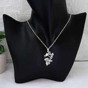 May include: A silver-colored dragon pendant necklace. The dragon has intricate details with blue and green accents. The necklace is displayed on a black mannequin.