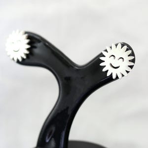 May include: A black, glossy, sewing tracing wheel with white, daisy-shaped ends, each featuring a smiling face. The wheel's handle has a curved, ergonomic design. The background is a soft, neutral color.