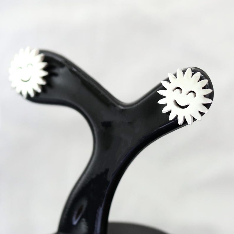 May include: A black, glossy, sewing tracing wheel with white, daisy-shaped ends, each featuring a smiling face. The wheel's handle has a curved, ergonomic design. The background is a soft, neutral color.