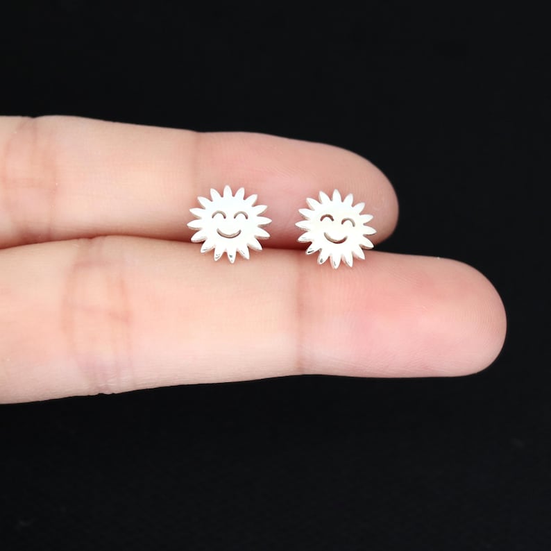 May include: A pair of small, silver-colored stud earrings. Each earring is shaped like a smiling sun with spiky rays. The earrings are held between fingers against a dark background.