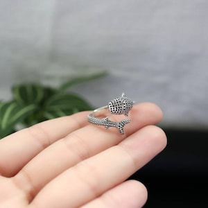 K&ouml;nnte beinhalten: Ein silberfarbener Ring in Form eines Walhais. Der Ring umschlie&szlig;t den Finger, wobei der K&ouml;rper und die Schwanzflosse des Walhais das Design bilden. Der K&ouml;rper des Walhais ist mit kleinen, dunklen Punkten bedeckt.
