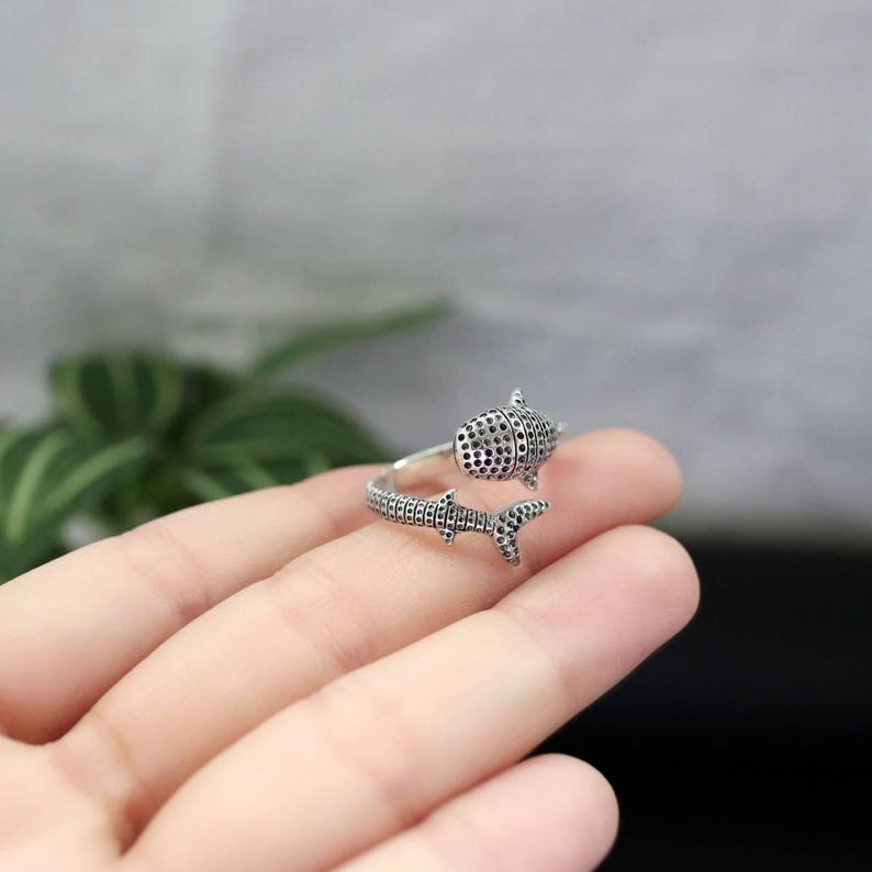 K&ouml;nnte beinhalten: Ein silberfarbener Ring in Form eines Walhais. Der Ring umschlie&szlig;t den Finger, wobei der K&ouml;rper und die Schwanzflosse des Walhais das Design bilden. Der K&ouml;rper des Walhais ist mit kleinen, dunklen Punkten bedeckt.
