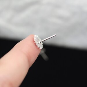 May include: A close-up of a silver-colored stud earring with a floral design. The earring is held between two fingers, with the post extending away from the flower-shaped front. The background is blurred, with a mix of light and dark tones.