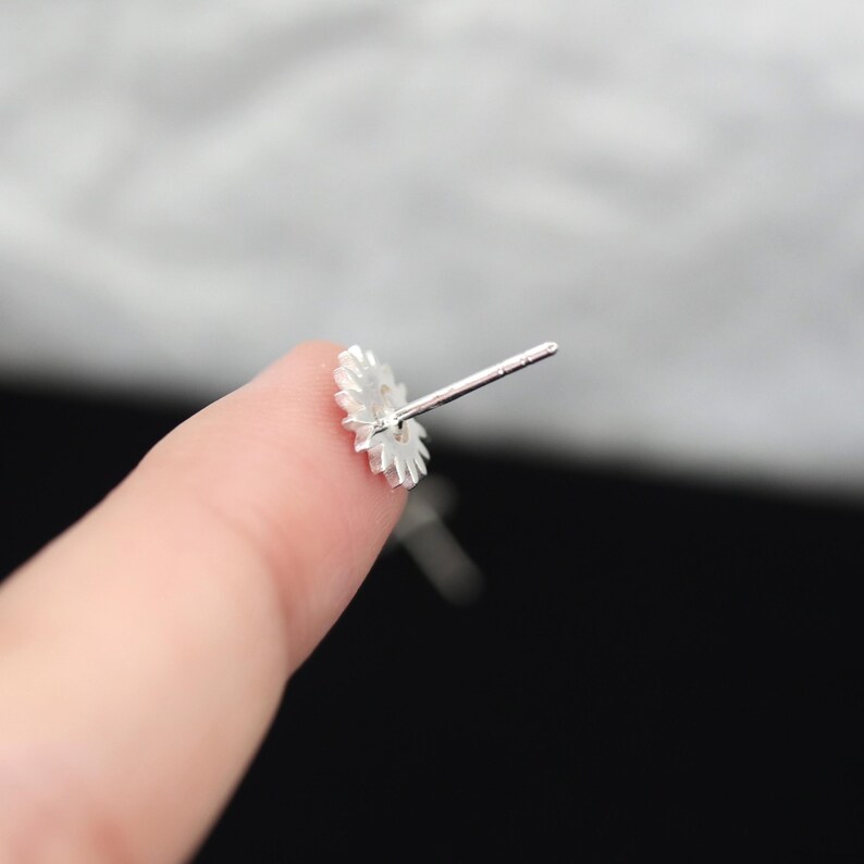 May include: A close-up of a silver-colored stud earring with a floral design. The earring is held between two fingers, with the post extending away from the flower-shaped front. The background is blurred, with a mix of light and dark tones.