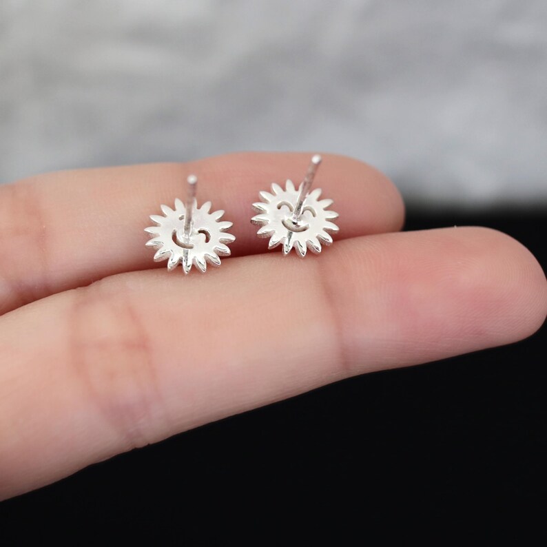 May include: Two silver-colored stud earrings. Each earring is shaped like a sun with a smiling face in the center. The sun's rays are pointed and evenly spaced around the face. The earrings are small and delicate.
