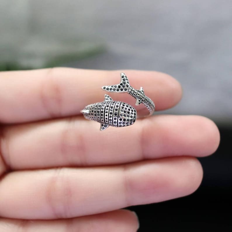 K&ouml;nnte beinhalten: Ein silberner Ring in Form eines Walhais. Der Ring hat ein detailliertes Design mit kleinen, dunklen Akzenten. Der Schwanz des Hais umschlingt den Finger und schafft ein einzigartiges und auff&auml;lliges Schmuckst&uuml;ck.