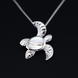 May include: A silver-colored turtle pendant necklace with a white shell inlay. The turtle has detailed flipper and head designs, and hangs from a delicate silver chain. The background is black.