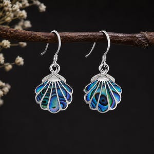 May include: A pair of silver drop earrings featuring a seashell-shaped pendant inlaid with iridescent blue and green abalone shell. The earrings have a polished silver finish and are suspended from simple silver hooks for a secure fit.