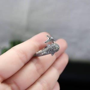 K&ouml;nnte beinhalten: Ein silberfarbener Ring in Form eines Walhais. Der Ring hat ein detailliertes Design mit kleinen schwarzen Akzenten, wodurch ein strukturiertes Aussehen entsteht. Der Schwanz und der K&ouml;rper des Walhais sind sichtbar.