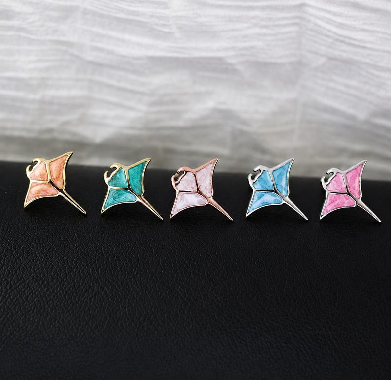 May include: Five stingray-shaped pins arranged in a row. Each pin features a metallic outline filled with marbled enamel in distinct colors: peach, teal, light pink, light blue, and bright pink. The pins have a pointed tail and wing-like fins.