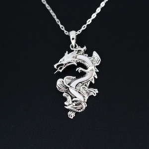 May include: A silver dragon pendant necklace. The pendant features a detailed dragon design with a white inlay, suspended from a delicate chain. The dragon is depicted with open mouth and intricate scales. The necklace is displayed against a black background.