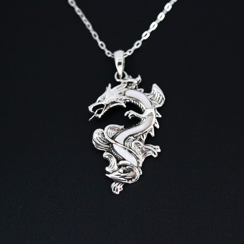 May include: A silver dragon pendant necklace. The pendant features a detailed dragon design with a white inlay, suspended from a delicate chain. The dragon is depicted with open mouth and intricate scales. The necklace is displayed against a black background.