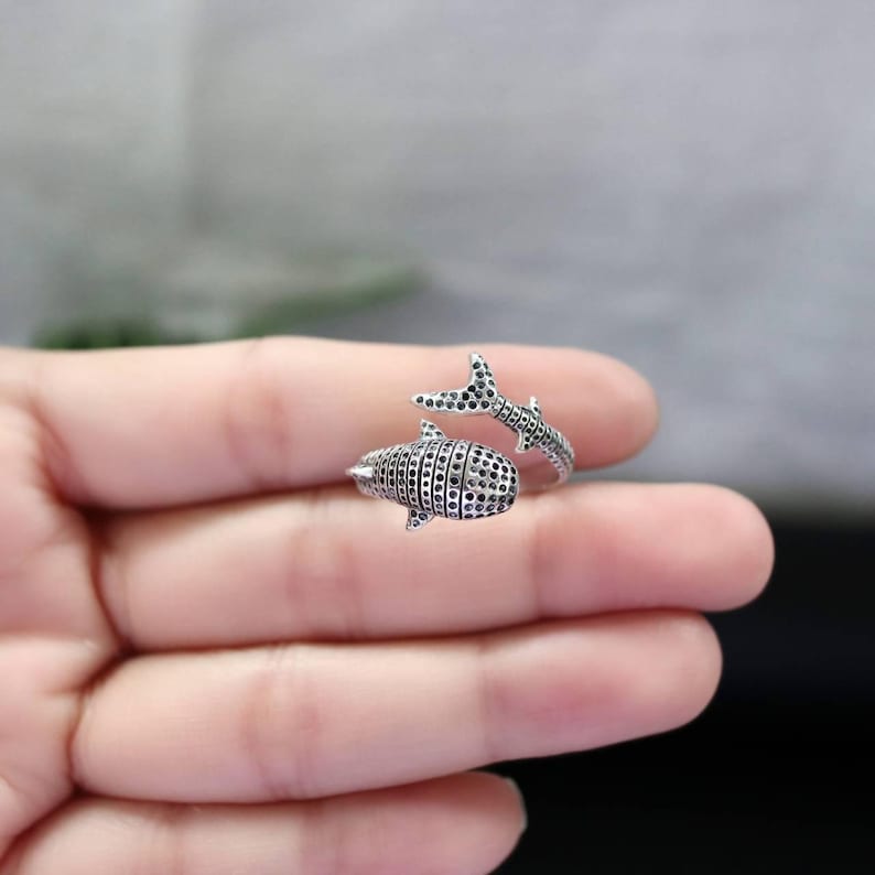 K&ouml;nnte beinhalten: Ein silberfarbener Ring in Form eines Walhais. Der Ring hat ein detailliertes Design mit einem strukturierten K&ouml;rper und einem Schwanz, der sich um den Finger wickelt. Der K&ouml;rper des Walhais ist mit kleinen, dunklen Punkten bedeckt.
