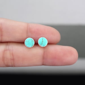 May include: A pair of round turquoise stud earrings with a silver setting. The earrings have a bright, robin's egg blue color with natural veining. The earrings are small and delicate, perfect for everyday wear.