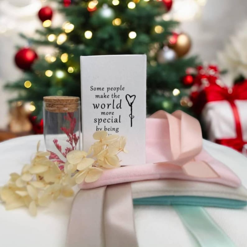 May include: A white rectangular card with black text reading Some people make the world more special by being in it, next to a small glass jar with a cork lid containing red dried flowers, pale yellow dried petals, and folded fabric ribbons.