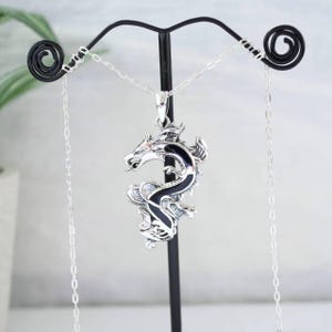 May include: A silver dragon pendant with black enamel detailing hangs from a delicate chain. The dragon is intricately designed, with detailed scales and a fierce expression. The pendant is displayed on a black metal stand against a soft background.