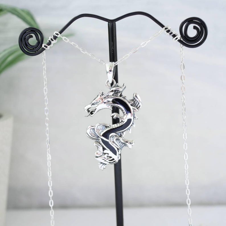 May include: A silver dragon pendant with black enamel detailing hangs from a delicate chain. The dragon is intricately designed, with detailed scales and a fierce expression. The pendant is displayed on a black metal stand against a soft background.
